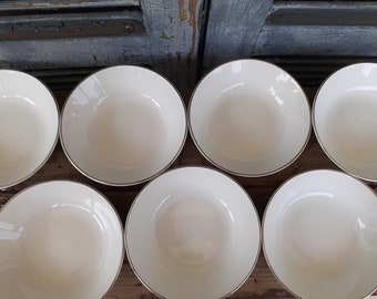 Puede incluir: Siete cuencos de cerámica blanca con un borde dorado. Los cuencos están dispuestos en semicírculo sobre una superficie de madera.
