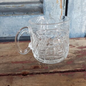 May include: A clear glass mug featuring a molded, three-dimensional relief design of the Batman logo and cityscape on its exterior surface. The mug has a smooth, curved handle and a circular base, showcasing a transparent finish throughout the piece.