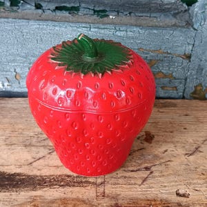 May include: A bright red strawberry-shaped lidded container with a textured surface featuring small indentations. The lid is topped with a green stem and leaf design. The object is made of a glossy material and has a rounded, bulbous shape.