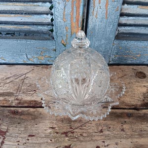 May include: A clear, cut-glass butter dish with a domed lid and starburst base. The dish features intricate floral and geometric patterns. The lid has a small, decorative knob. The dish is displayed on a rustic wooden surface.