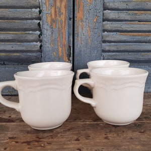May include: Four white ceramic mugs with a scalloped design. The mugs are arranged in a row on a wooden surface.