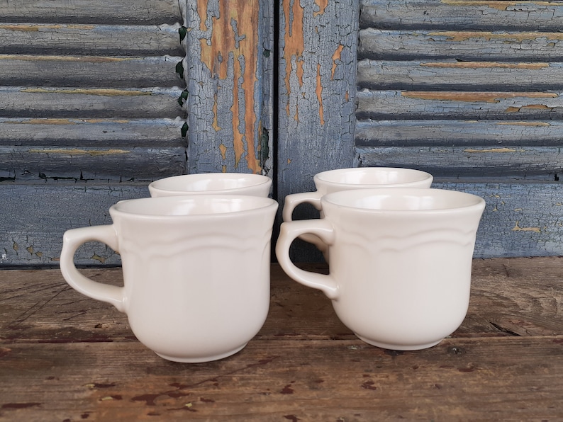 May include: Four white ceramic mugs with a scalloped design. The mugs are arranged in a row on a wooden surface.