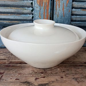 May include: A white ceramic serving bowl with a lid. The bowl has a rounded shape and a small, cylindrical handle on the lid. The bowl is sitting on a wooden surface.