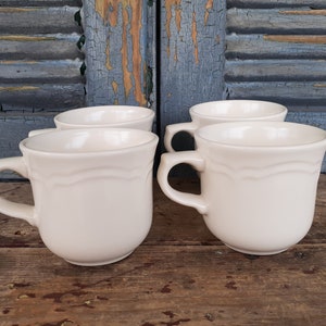 May include: Four off-white ceramic mugs with a simple design. The mugs have a rounded shape and a single handle.