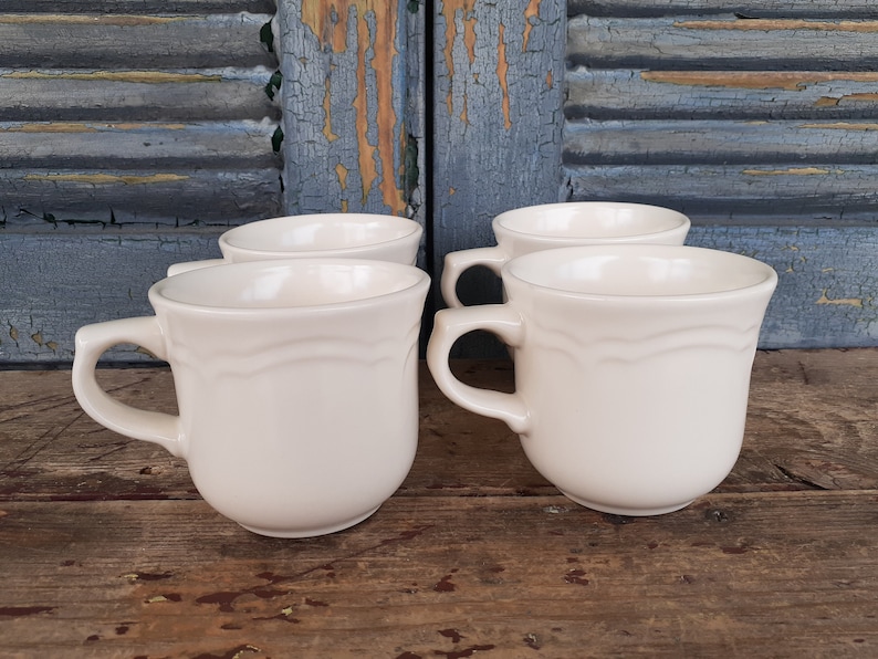 May include: Four off-white ceramic mugs with a simple design. The mugs have a rounded shape and a single handle.