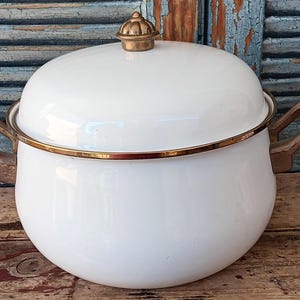 May include: A white enameled metal pot with a matching lid and a gold-toned metal knob. The pot features two curved gold-toned metal handles on the sides and a thin gold-toned metal rim around the top edge of the base and the lid.