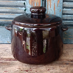 May include: A dark brown ceramic pot with a lid. The pot has two small handles and a rounded lid with a knob. The pot is sitting on a wooden surface.
