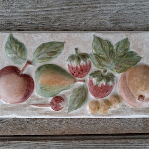 May include: Rectangular ceramic tile with a relief design of various fruits. The fruits include a red apple, a green pear, red strawberries, and a peach. Green leaves and small yellow grapes are also present. The tile has a cream-colored background.