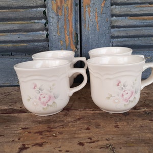 May include: Four white ceramic mugs with a floral design featuring pink roses and green leaves. The mugs have a scalloped rim and a handle.