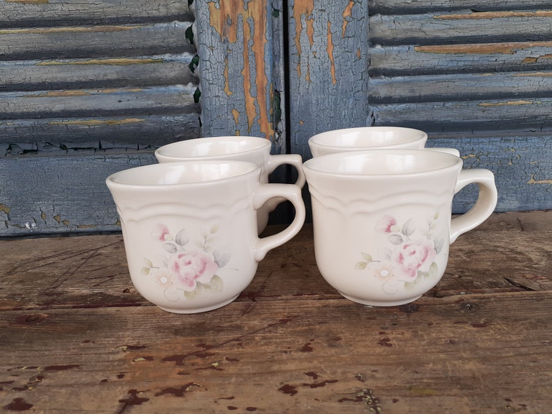 May include: Four white ceramic mugs with a floral design featuring pink roses and green leaves. The mugs have a scalloped rim and a handle.