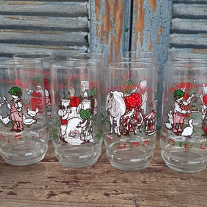 May include: Set of six clear glass drinking glasses with a Christmas theme. Each glass features a different illustration of a child in a red dress and a green hat, with a different verse of the song "The Twelve Days of Christmas" printed below the illustration.