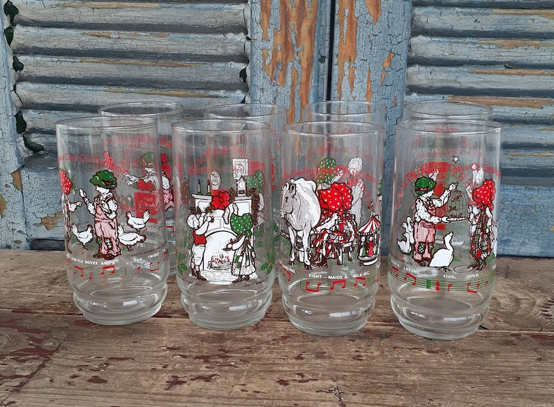 May include: Set of six clear glass drinking glasses with a Christmas theme. Each glass features a different illustration of a child in a red dress and a green hat, with a different verse of the song "The Twelve Days of Christmas" printed below the illustration.