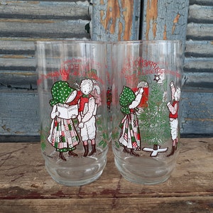 May include: Two vintage glass tumblers with Christmas scenes. One features two children singing carols, the other shows two children decorating a Christmas tree. The text on the glasses reads "Twelve Days Of Christmas" and "A Little Christmas Shop & Little Christmas Tree".