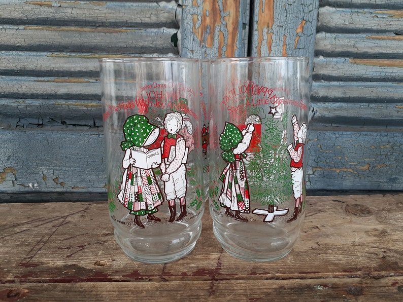 May include: Two vintage glass tumblers with Christmas scenes. One features two children singing carols, the other shows two children decorating a Christmas tree. The text on the glasses reads "Twelve Days Of Christmas" and "A Little Christmas Shop & Little Christmas Tree".