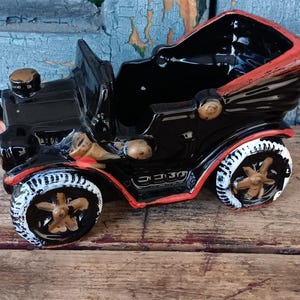 May include: A vintage black ceramic car-shaped planter with red and gold accents. The planter features white-rimmed wheels with gold spokes. The car has a classic, antique design, perfect for small plants or decorative use.