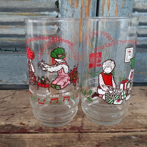 May include: Two clear glass Christmas themed drinking glasses. The glass on the left has a red and green illustration of the "Twelve Days of Christmas" with the text "Eleven Pipers Piping". The glass on the right has a red and green illustration of a girl wrapping presents with the text "Wrap Each Christmas Gift".