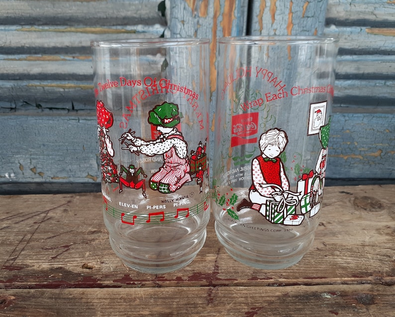 May include: Two clear glass Christmas themed drinking glasses. The glass on the left has a red and green illustration of the "Twelve Days of Christmas" with the text "Eleven Pipers Piping". The glass on the right has a red and green illustration of a girl wrapping presents with the text "Wrap Each Christmas Gift".