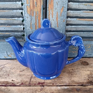 May include: A cobalt blue ceramic teapot with a matching lid. The teapot has a curved spout and handle, and a floral design on the handle. The teapot is sitting on a wooden surface.