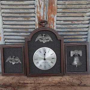 May include: A wooden wall clock with a silver face and black Roman numeral hour markers. The clock is surrounded by three decorative plaques featuring an eagle and a bell. The clock face reads "Seth Thomas".