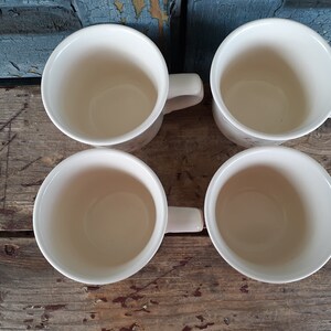 May include: Four white ceramic mugs with handles. The mugs are empty and sitting on a wooden surface.