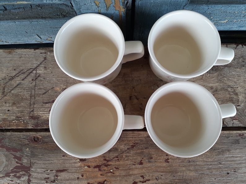 May include: Four white ceramic mugs with handles. The mugs are empty and sitting on a wooden surface.