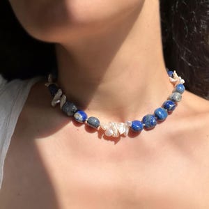 May include: A necklace featuring a strand of blue lapis lazuli and white pearl beads. The necklace is composed of irregularly shaped, polished lapis lazuli stones interspersed with smaller, white pearl accents. The necklace is worn around the neck.