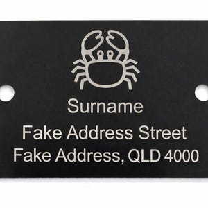 May include: Black rectangular sign featuring a crab emblem and the text "Surname", "Fake Address Street", and "Fake Address, QLD 4000". The sign has two mounting holes.