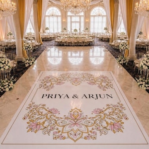 May include: A lavishly decorated ballroom, ideal for a wedding celebration. The dance floor is white, with the names "PRIYA & ARJUN" in the centre, framed by a pink and gold floral design. The room is lit by chandeliers and decorated with flowers.
