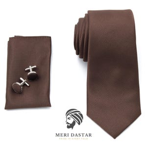 May include: A brown tie, pocket square, and cufflinks set. The tie and pocket square have a subtle ribbed texture. The cufflinks are round with a brown center and silver-colored metal. The set is displayed on a white background.