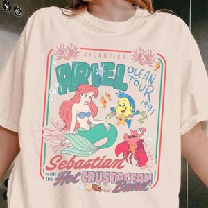 Pu&ograve; includere: T-shirt color crema con grafica colorata di Ariel, Flounder e Sebastian de La Sirenetta. Il design include il testo "Ariel Ocean Tour 1991" e "Sebastian with the Hot Crustacean Band."