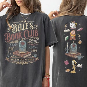 May include: Dark gray t-shirt featuring "Belle's Book Club" text and graphics from Beauty and the Beast. The front design includes a rose in a glass dome, books, and the text "Tale as old as Time". The back has additional character illustrations.