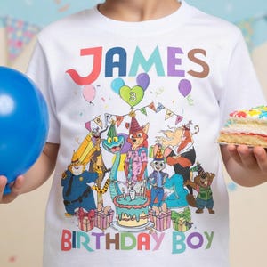 Pode incluir: Camiseta branca com o nome "JAMES" em letras coloridas e a mensagem "BIRTHDAY BOY". A camiseta apresenta uma ilustra&ccedil;&atilde;o de desenho animado de animais em volta de um bolo de anivers&aacute;rio. Um bal&atilde;o azul e um peda&ccedil;o de bolo tamb&eacute;m s&atilde;o vis&iacute;veis.