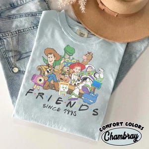 Puede incluir: Una camiseta azul claro con un estampado gr&aacute;fico de varios personajes de juguetes animados sentados juntos en un sof&aacute;. Debajo de los personajes, el texto "FRIENDS SINCE 1995" aparece en letras negras. La camiseta se muestra junto a un sombrero beige y vaqueros.