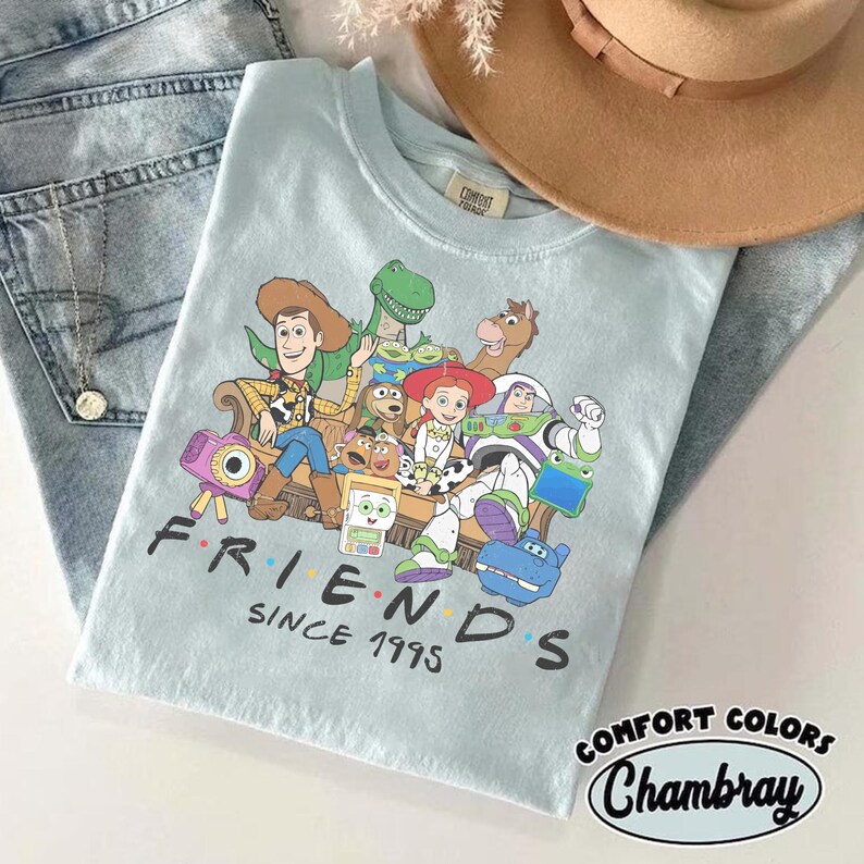 Puede incluir: Una camiseta azul claro con un estampado gr&aacute;fico de varios personajes de juguetes animados sentados juntos en un sof&aacute;. Debajo de los personajes, el texto "FRIENDS SINCE 1995" aparece en letras negras. La camiseta se muestra junto a un sombrero beige y vaqueros.