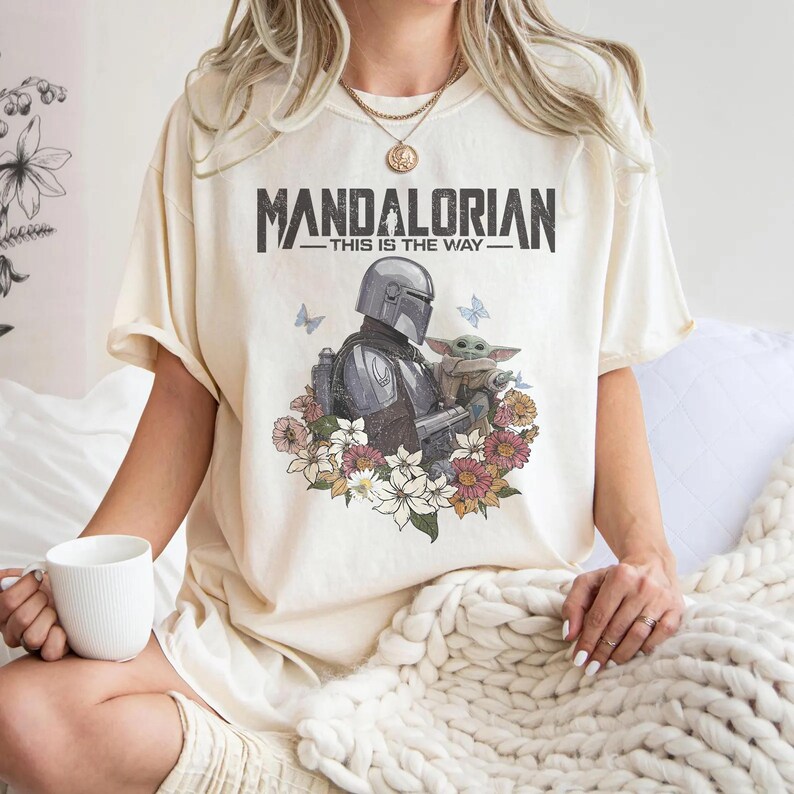 May include: Cream-coloured t-shirt with a graphic of the Mandalorian and Grogu surrounded by flowers and butterflies. The text "MANDALORIAN - THIS IS THE WAY" is printed above the graphic. The shirt has short sleeves.