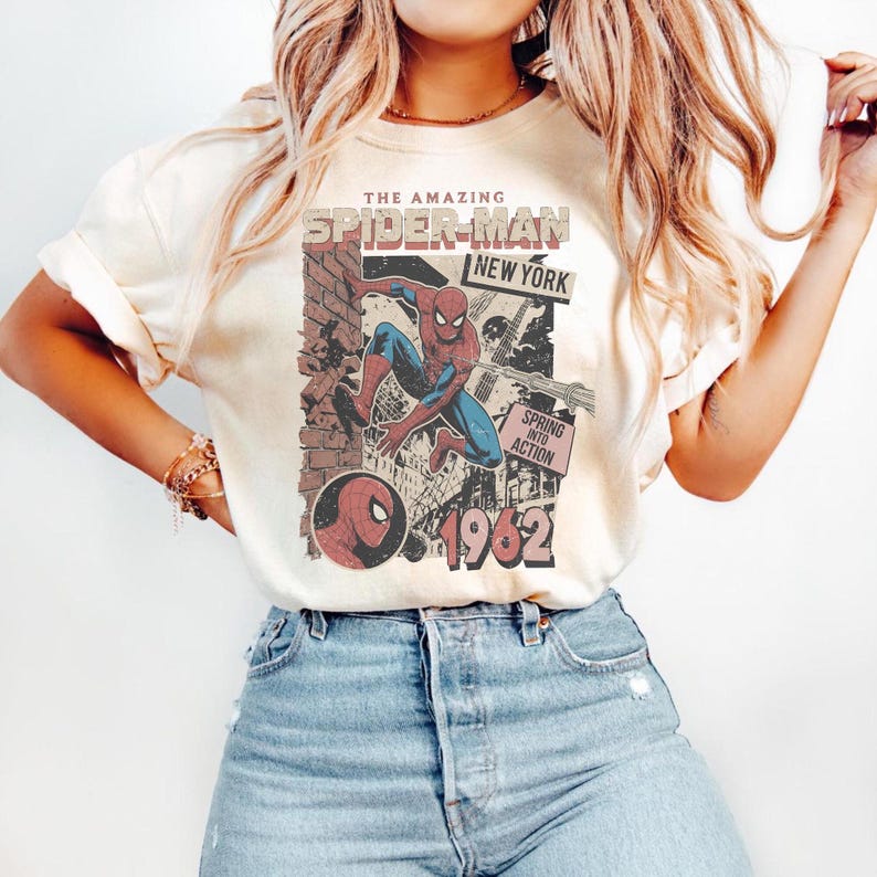 Op de afbeelding: Cr&egrave;mekleurig T-shirt met een vintage Spider-Man-afbeelding. Het ontwerp bevat de tekst "THE AMAZING SPIDER-MAN NEW YORK" en "1962". De afbeelding toont Spider-Man in actie, met een bakstenen muur en stadsachtergrond.