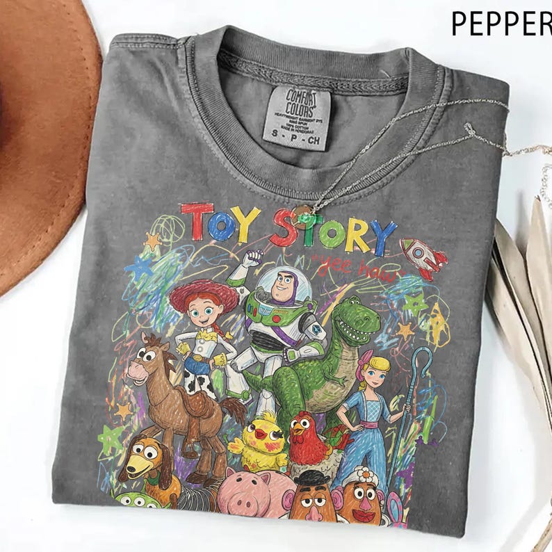 K&ouml;nnte beinhalten: Ein anthrazitfarbenes Kurzarm-T-Shirt mit einem bunten Grafikdruck der Toy Story-Figuren, darunter Woody, Buzz Lightyear, Jessie, Rex und Bo Peep. Das Design enth&auml;lt krakelige Buntstift-Akzente und das Toy Story-Logo auf der Vorderseite.