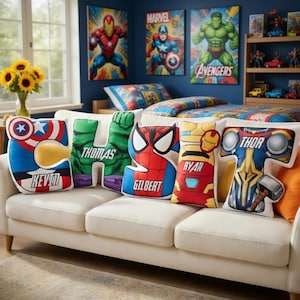 May include: A white sofa displays five superhero-themed pillows shaped like letters. The pillows feature Captain America, Hulk, Spider-Man, Iron Man, and Thor designs, with names on each. Marvel artwork and bedding are visible in the background.
