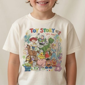 K&ouml;nnte beinhalten: Ein cremefarbenes Kurzarm-T-Shirt mit einer bunten Buntstiftzeichnung der Toy Story-Figuren, darunter Buzz Lightyear, Woody, Jessie, Rex und andere. Der Text TOY STORY und yee haw ist &uuml;ber der Charakterillustration aufgedruckt.