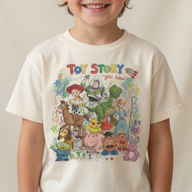 K&ouml;nnte beinhalten: Ein cremefarbenes Kurzarm-T-Shirt mit einer bunten Buntstiftzeichnung der Toy Story-Figuren, darunter Buzz Lightyear, Woody, Jessie, Rex und andere. Der Text TOY STORY und yee haw ist &uuml;ber der Charakterillustration aufgedruckt.