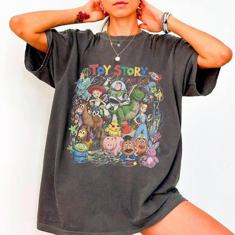 K&ouml;nnte beinhalten: Ein anthrazitfarbenes Kurzarm-T-Shirt mit einem bunten Grafikdruck der Toy Story-Figuren, darunter Buzz Lightyear, Woody, Jessie und Rex, sowie dem Schriftzug TOY STORY in fetten, mehrfarbigen Buchstaben im oberen Bereich.