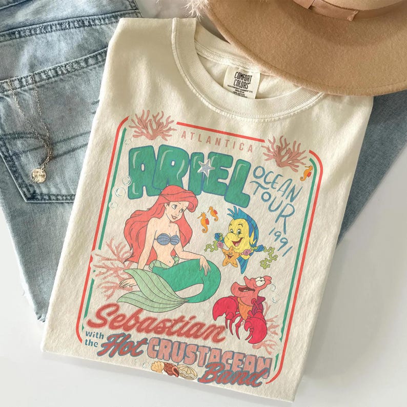 Pu&ograve; includere: T-shirt color crema con una grafica di Ariel de La Sirenetta, con la scritta "Atlantica Ariel Ocean Tour 1991". Il design include illustrazioni di Ariel, Flounder, Sebastian e altre creature marine. La maglietta &egrave; piegata con un cappello e jeans.