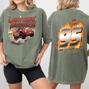Op de afbeelding: Olijfgroen T-shirt met Lightning McQueen uit de film Cars. De afbeelding op de voorkant toont McQueen in de race met de tekst "Lightning McQueen" erboven. De achterkant van het shirt toont het nummer "95" in vlammen met de tekst "Lightning McQueen".