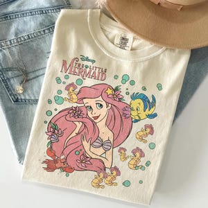 Puede incluir: Una camiseta de color crema con un estampado gr&aacute;fico de Ariel de La Sirenita de Disney, rodeada por Flounder, Sebasti&aacute;n y varios caballitos de mar. El dise&ntilde;o incluye burbujas verde azulado y flores rosas, con el t&iacute;tulo de la pel&iacute;cula arriba.