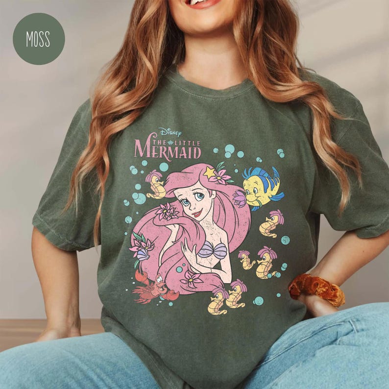 Puede incluir: Una camiseta de manga corta color verde musgo con un estampado gr&aacute;fico de Ariel de La Sirenita con el pelo rosa, rodeada de Flounder, Sebasti&aacute;n y caballitos de mar. El texto Disney The Little Mermaid aparece impreso sobre la ilustraci&oacute;n.