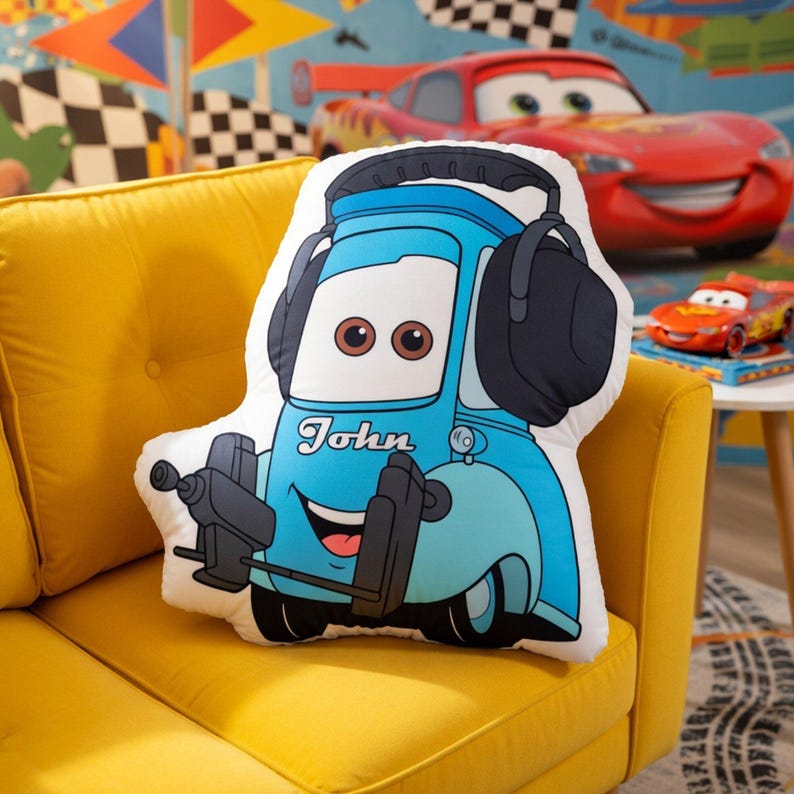 May include: A shaped throw pillow featuring a cartoon character resembling a blue tow truck wearing large black headphones. The character has brown eyes, a wide smile, and the name John printed in white script across the front hood area.