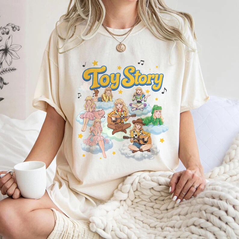 May include: A cream-colored short-sleeve t-shirt featuring a graphic print with the text Toy Story in yellow letters. The design includes multiple illustrated characters sitting on clouds, musical notes, and yellow stars scattered around the text.