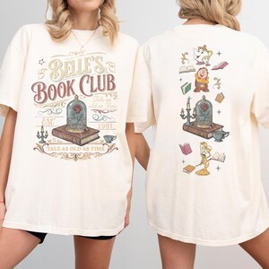 May include: Cream-colored t-shirt featuring the text "Belle's Book Club" with a rose in a glass dome graphic. The back of the shirt has illustrations of characters and objects from the Beauty and the Beast movie. The shirt has short sleeves.