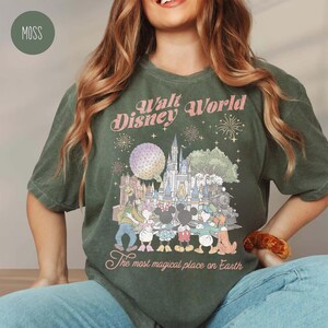 May include: Moss green t-shirt featuring a Walt Disney World graphic. The design includes the park's castle, characters, and the text "The most magical place on Earth". The shirt has a vintage, distressed look.