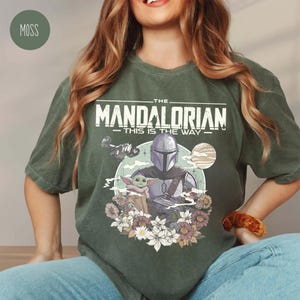 May include: A moss green short-sleeve t-shirt featuring a graphic print of The Mandalorian and Grogu. The design includes the text 'THE MANDALORIAN' and 'THIS IS THE WAY' above the characters, surrounded by a circular background and floral accents.